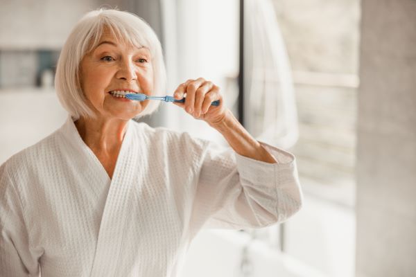 An older woman stands in front of a bathroom mirror and brushes her teeth